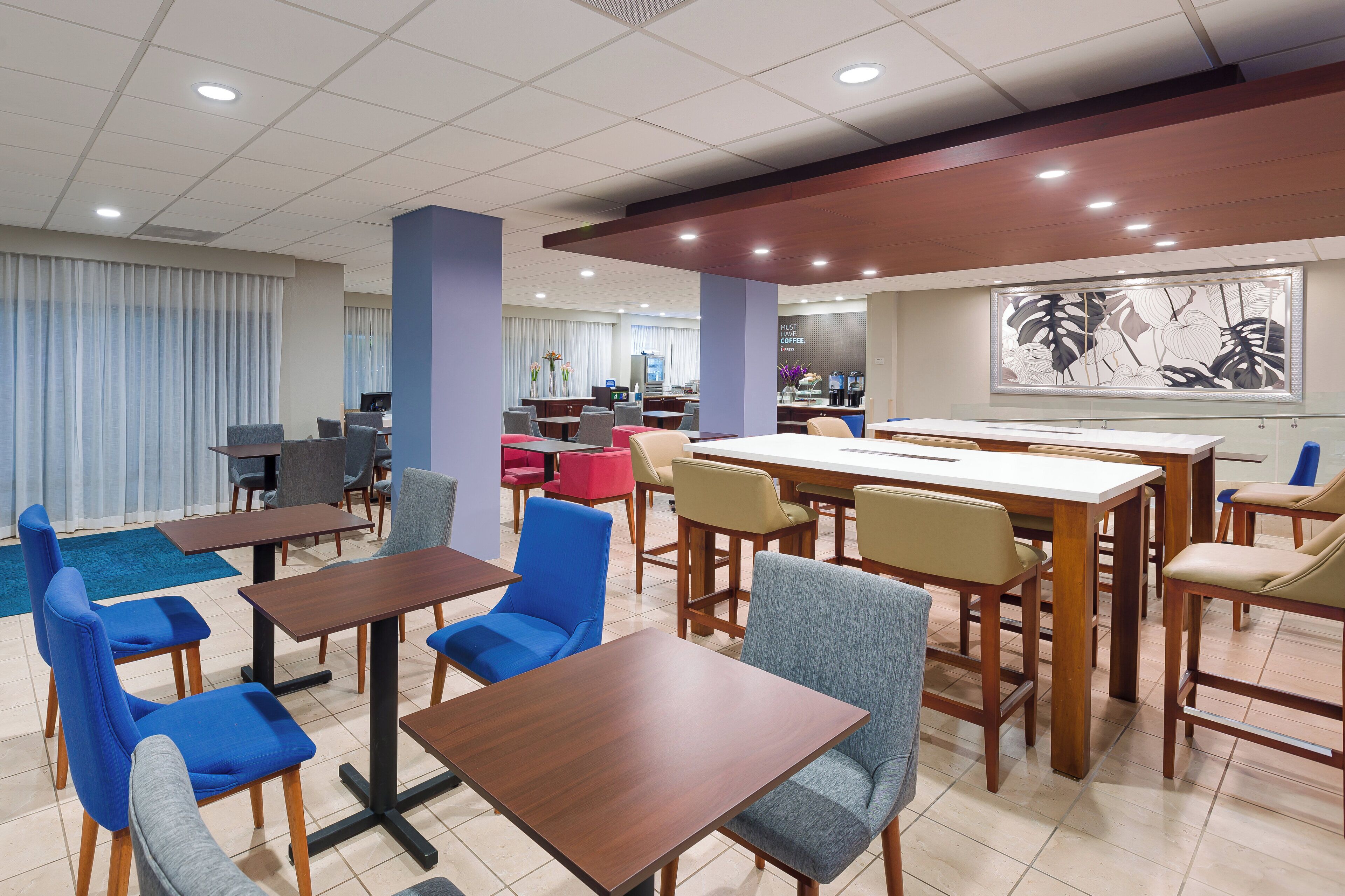 A hotel dining area with multiple tables and chairs, a coffee station, and a large abstract art piece on the wall.