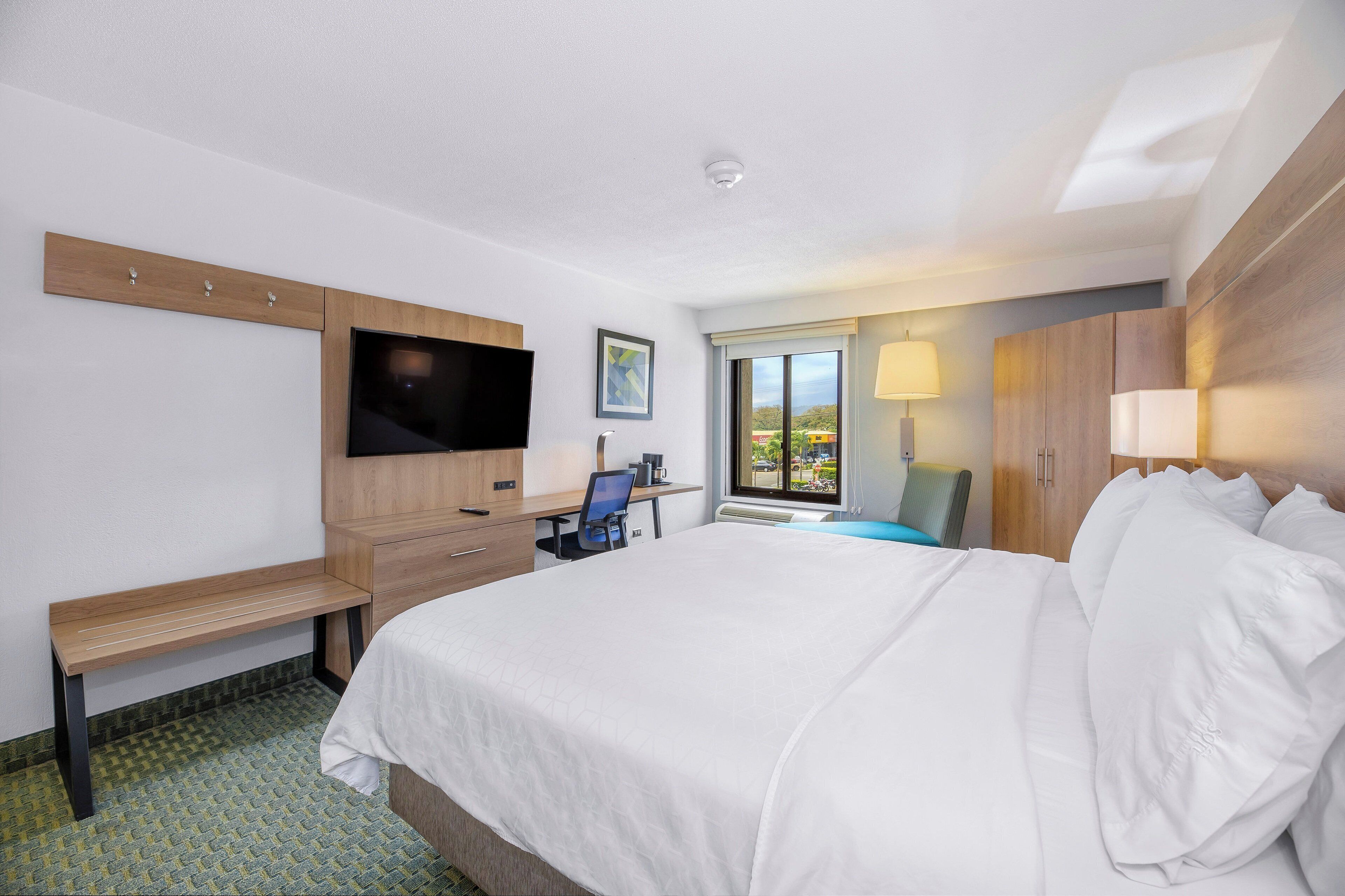 A hotel room with a large bed, a desk with a chair, a TV mounted on the wall, and a window with a view of the outside.
