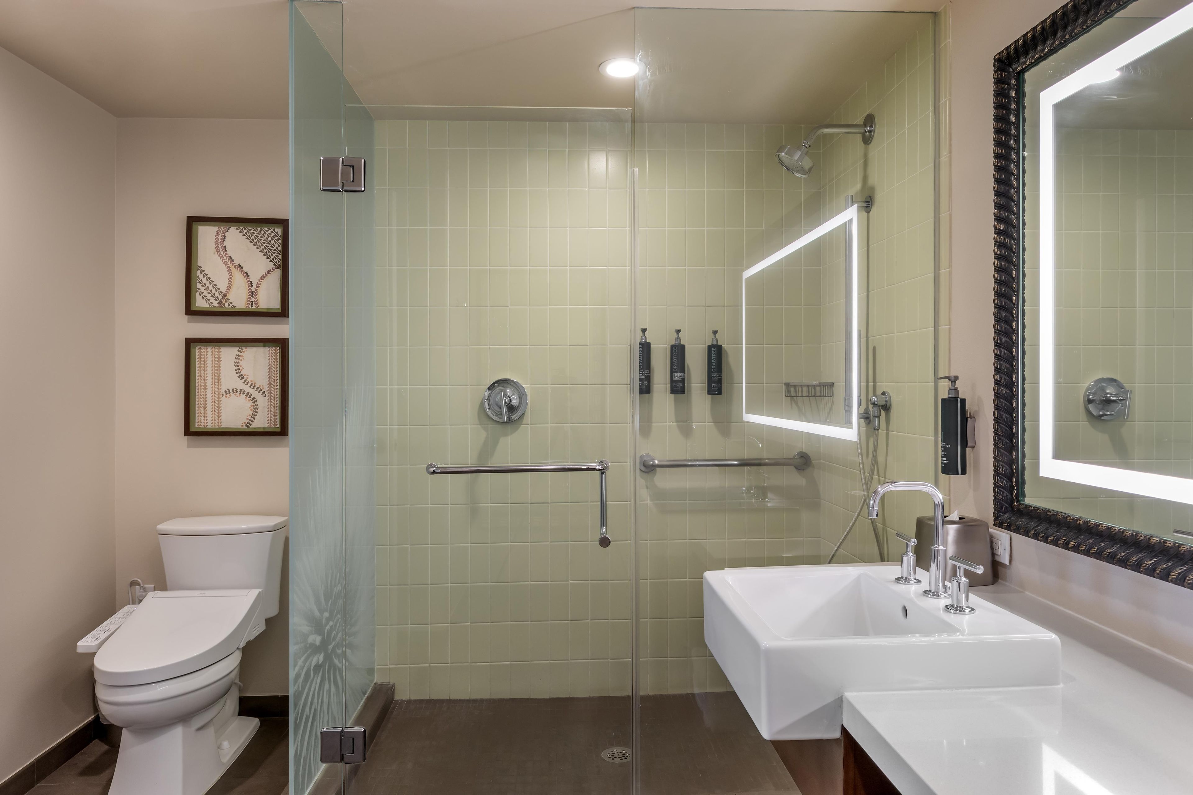 A bathroom with a toilet, a shower with glass doors, a sink with a mirror, and framed artwork on the wall.