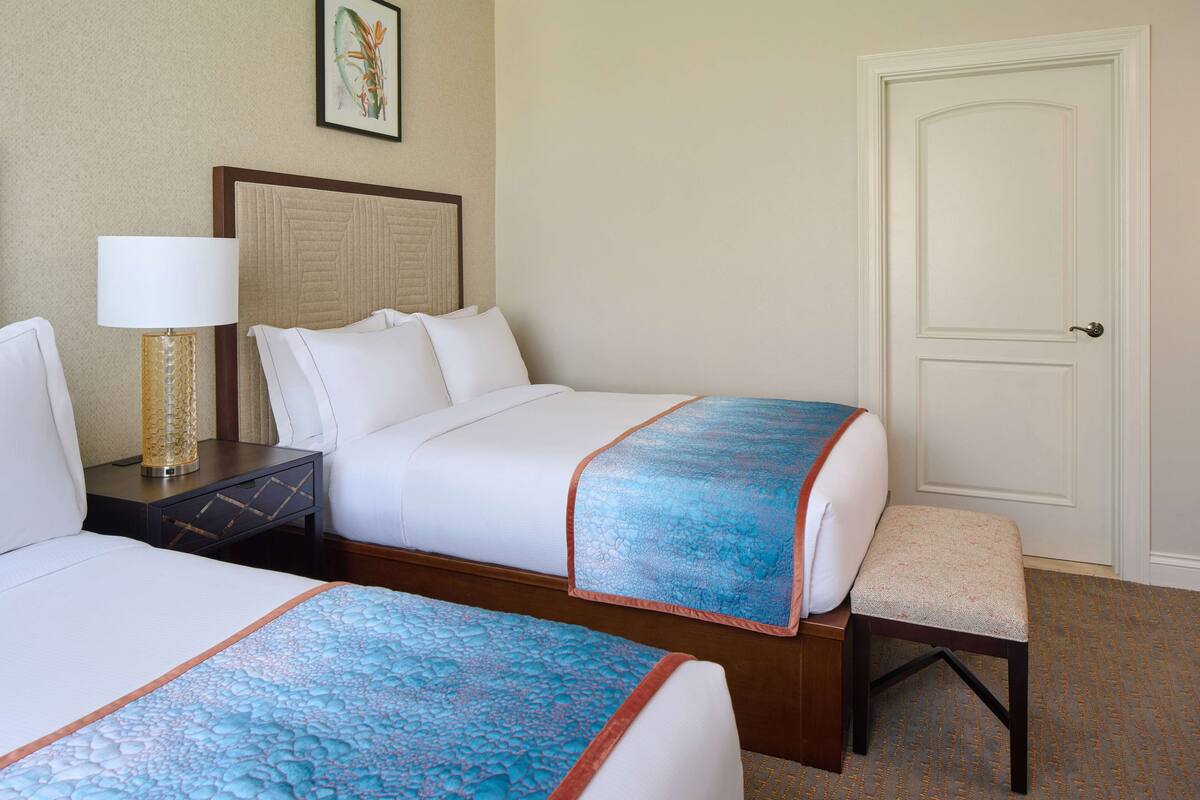 Une chambre d’hôtel avec deux lits, une table de chevet avec une lampe, un banc et une porte.