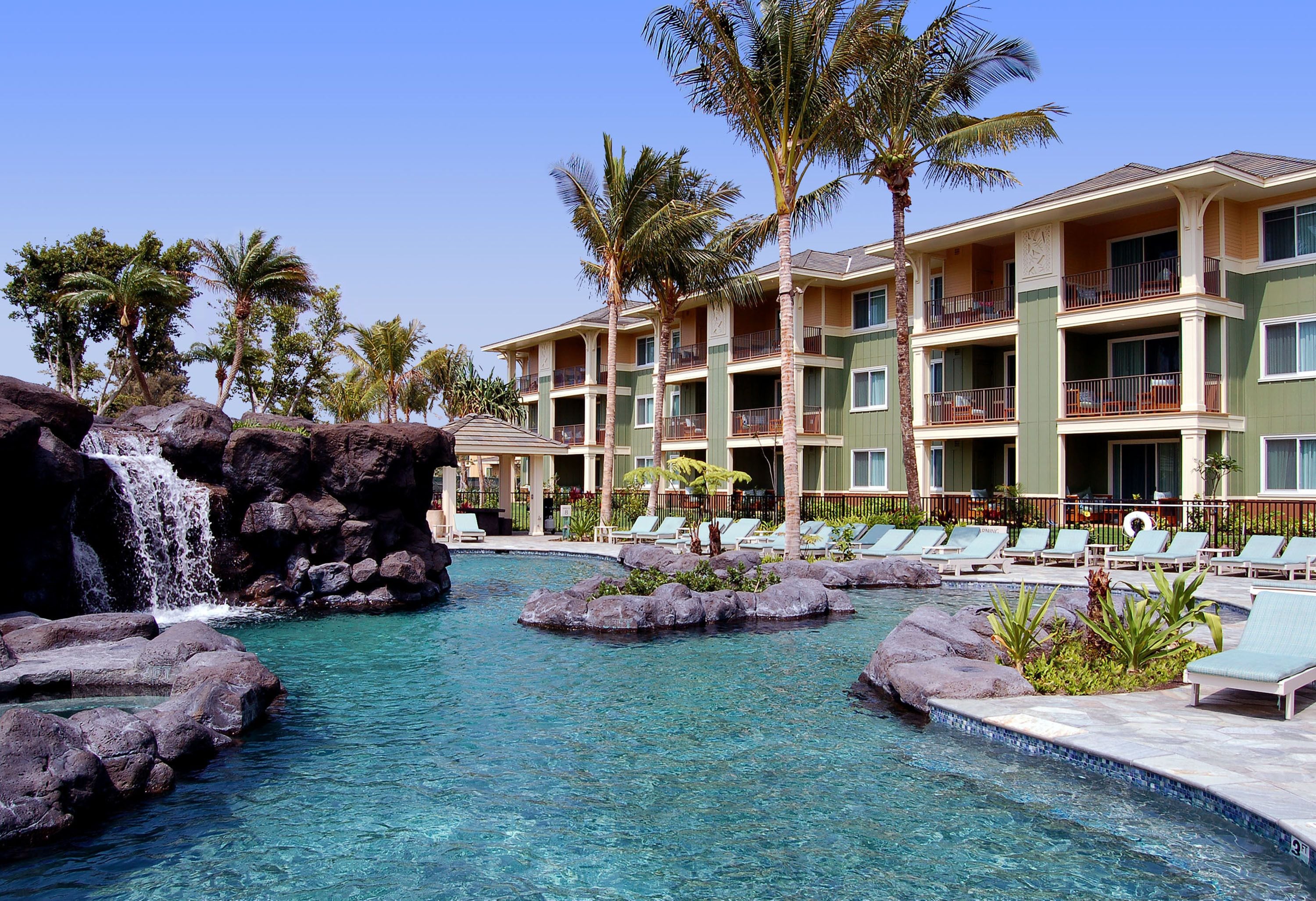 A hotel with a pool featuring a waterfall and rock formations, surrounded by palm trees and lounge chairs.