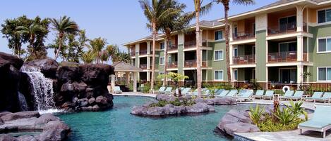 A hotel with a pool featuring a waterfall and rock formations, surrounded by palm trees and lounge chairs.