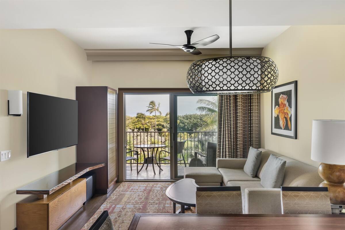 Une chambre d’hôtel moderne dotée d’une télévision à écran plat, d’un ventilateur de plafond, d’un coin repas et d’un balcon offrant une vue sur les palmiers.