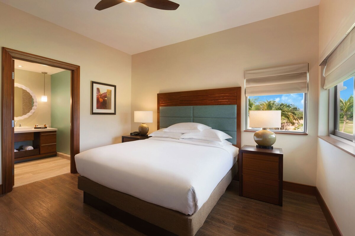 Une chambre d’hôtel comprenant un lit, des tables de chevet, une fenêtre avec vue, un ventilateur de plafond et une salle de bain avec un miroir et un lavabo.