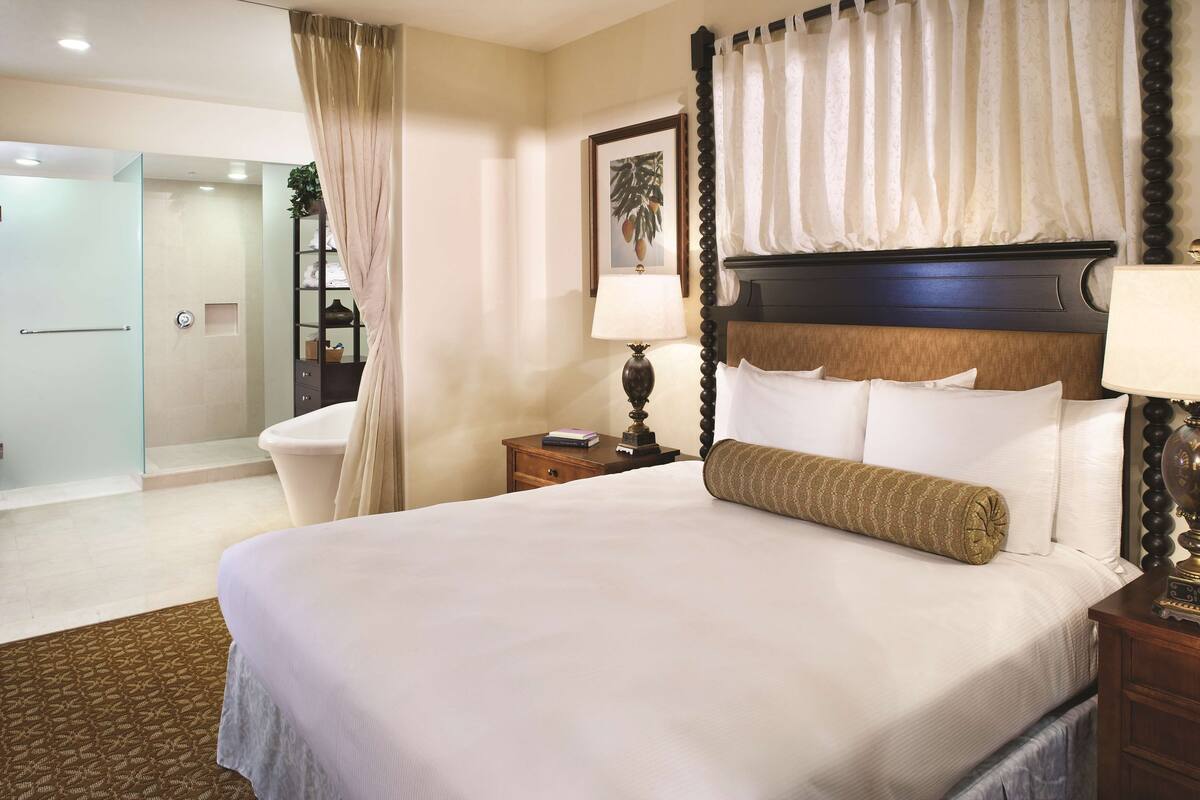 Une chambre d’hôtel avec un grand lit, une table de chevet avec une lampe, une salle de bain avec douche et une porte vitrée donnant sur une autre pièce.