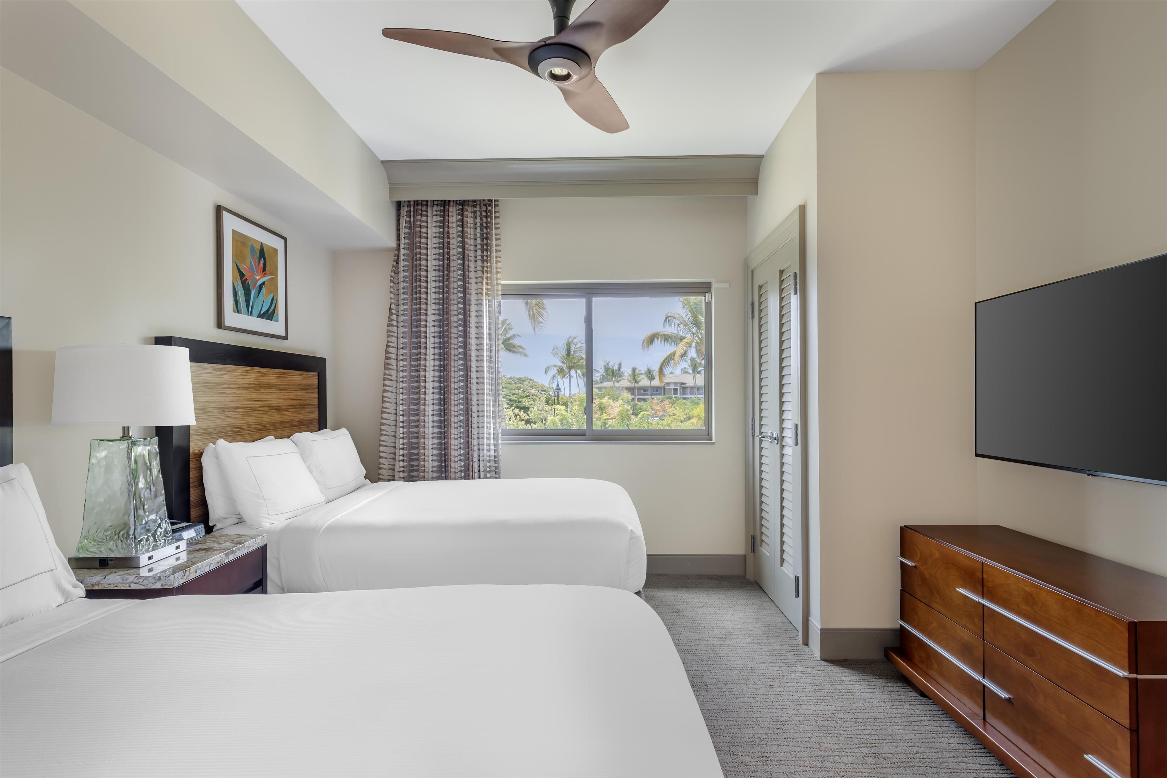 A hotel room with two beds, a ceiling fan, a flat-screen TV, a nightstand, a lamp, a dresser, and a window with a view of palm trees.