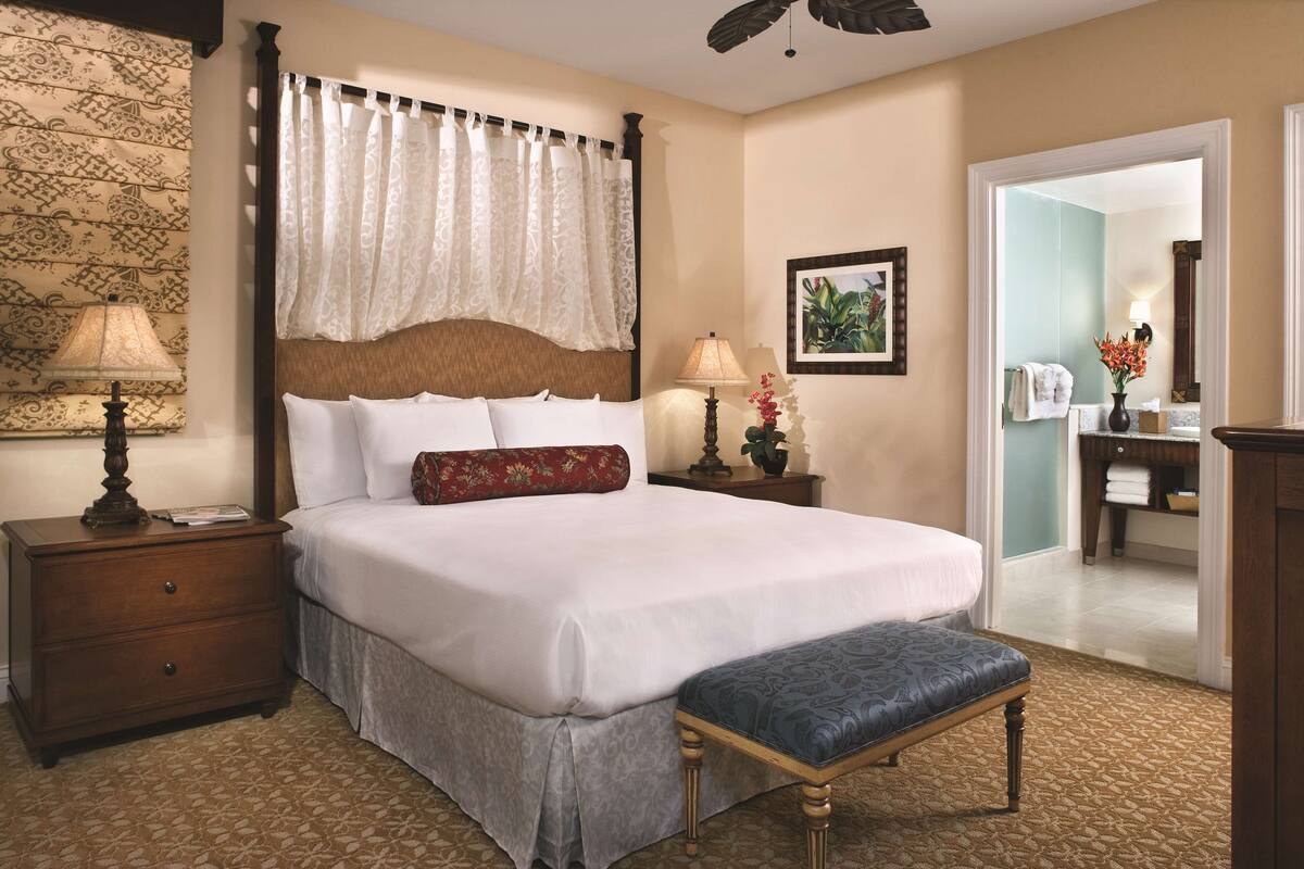 Une chambre d’hôtel avec un grand lit, une table de chevet en bois, un banc, une moquette à motifs, un mur orné d’un panneau décoratif et une salle de bain visible à travers une porte ouverte.