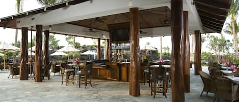 An outdoor bar area with stone flooring, wooden pillars, and wicker furniture.