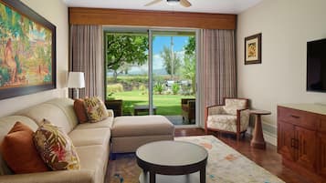 A hotel room with a sofa, armchair, round table, and a view of a garden through a sliding door.