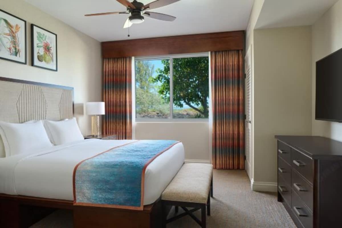 A hotel room with a bed, a bench, a TV, a ceiling fan, and a window with curtains.