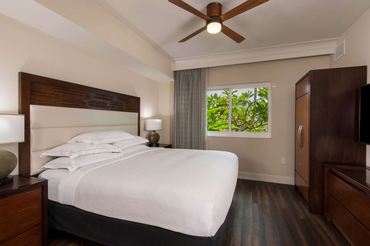 Une chambre d’hôtel avec un lit, un ventilateur de plafond, une fenêtre donnant sur un beau paysage, une télévision et des meubles en bois.
