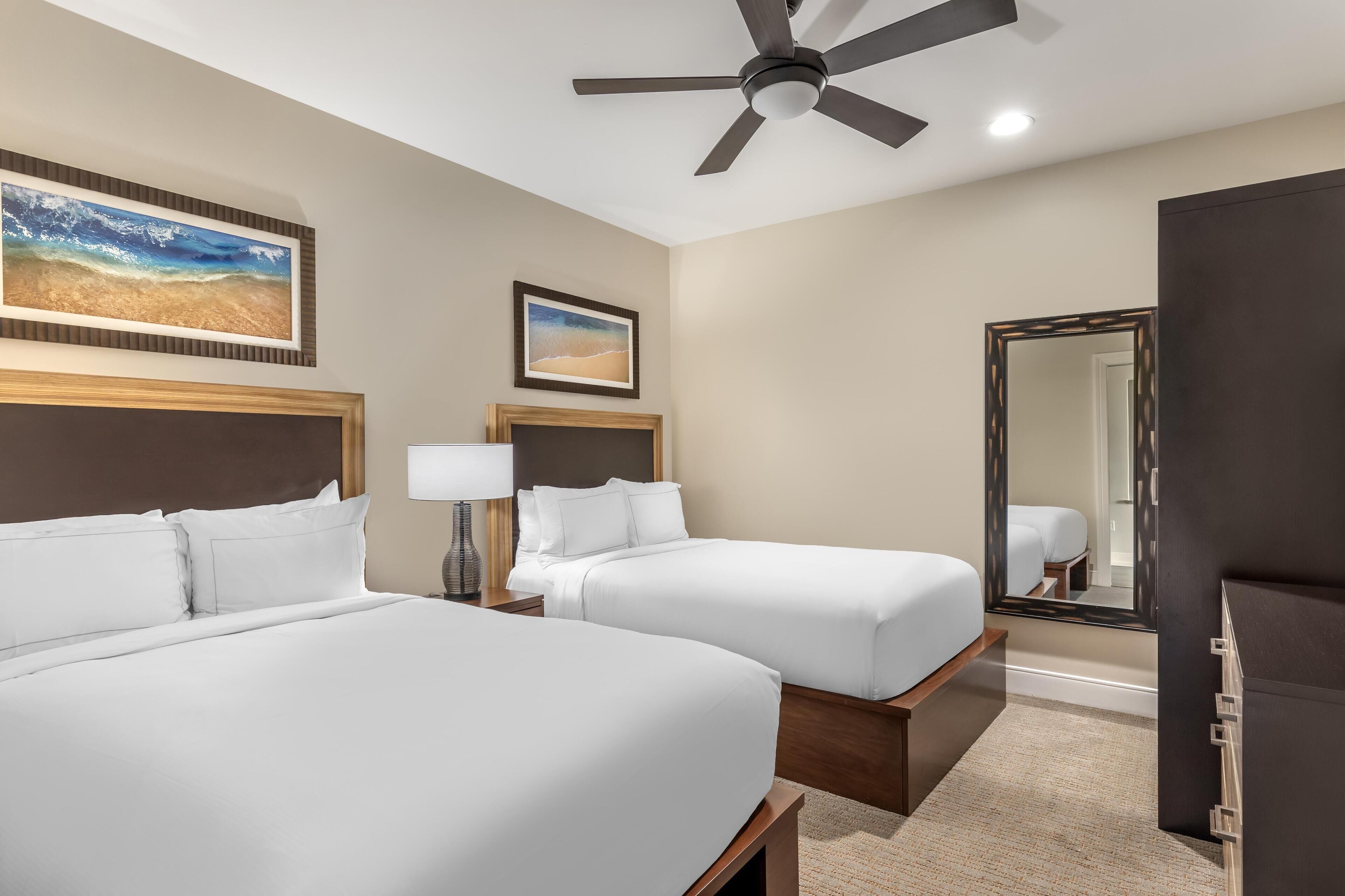 A hotel room with two beds, a ceiling fan, a large mirror, and artwork on the walls.