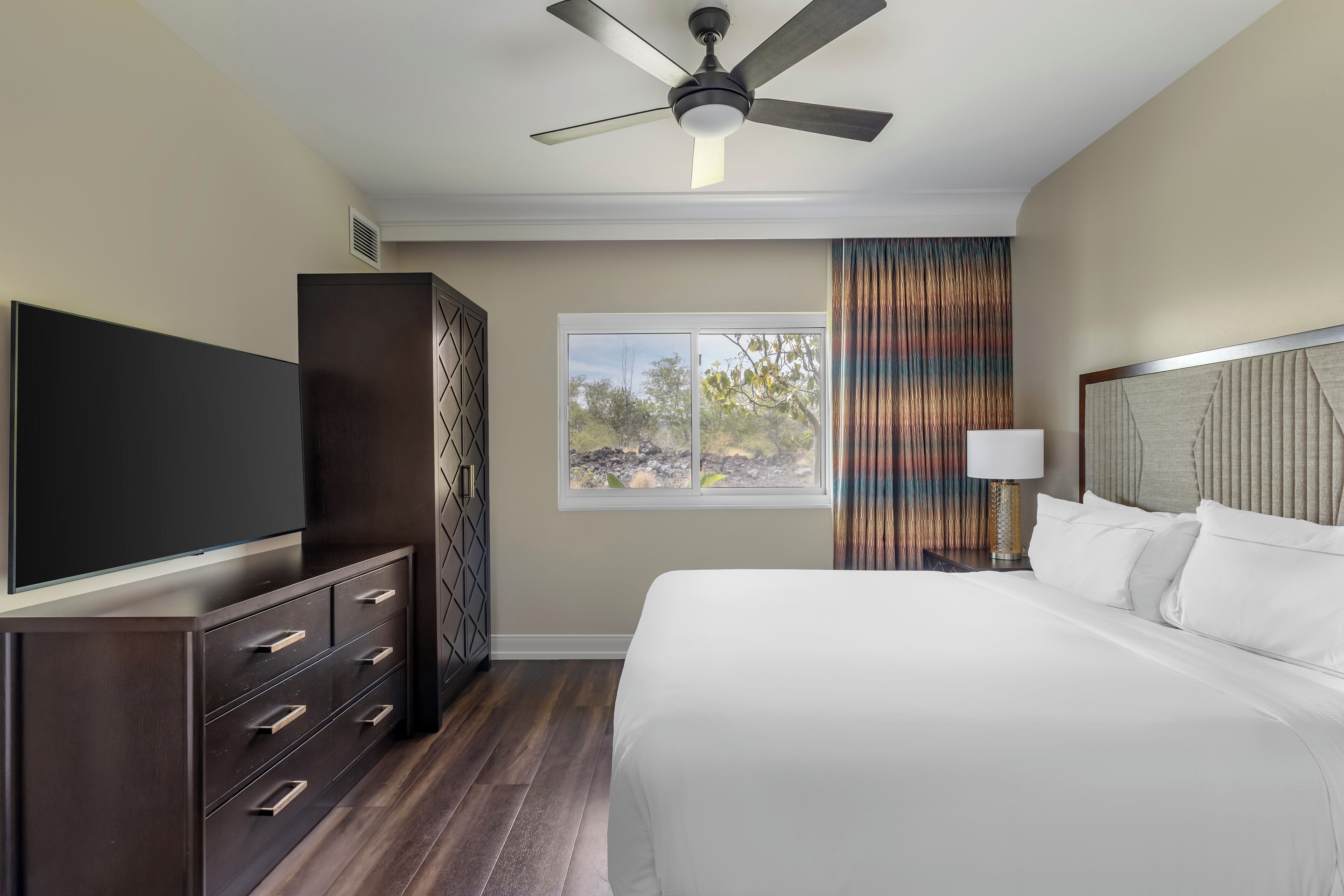 Una habitación de hotel con cama, televisor, armario, ventilador de techo, ventana con cortinas y mesita de noche con lámpara.