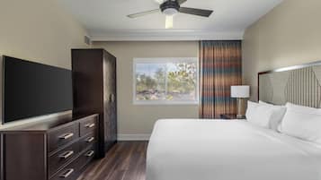 Una habitación de hotel con cama, televisor, armario, ventilador de techo, ventana con cortinas y mesita de noche con lámpara.