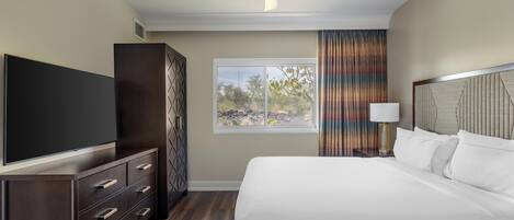 Una habitación de hotel con cama, televisor, armario, ventilador de techo, ventana con cortinas y mesita de noche con lámpara.