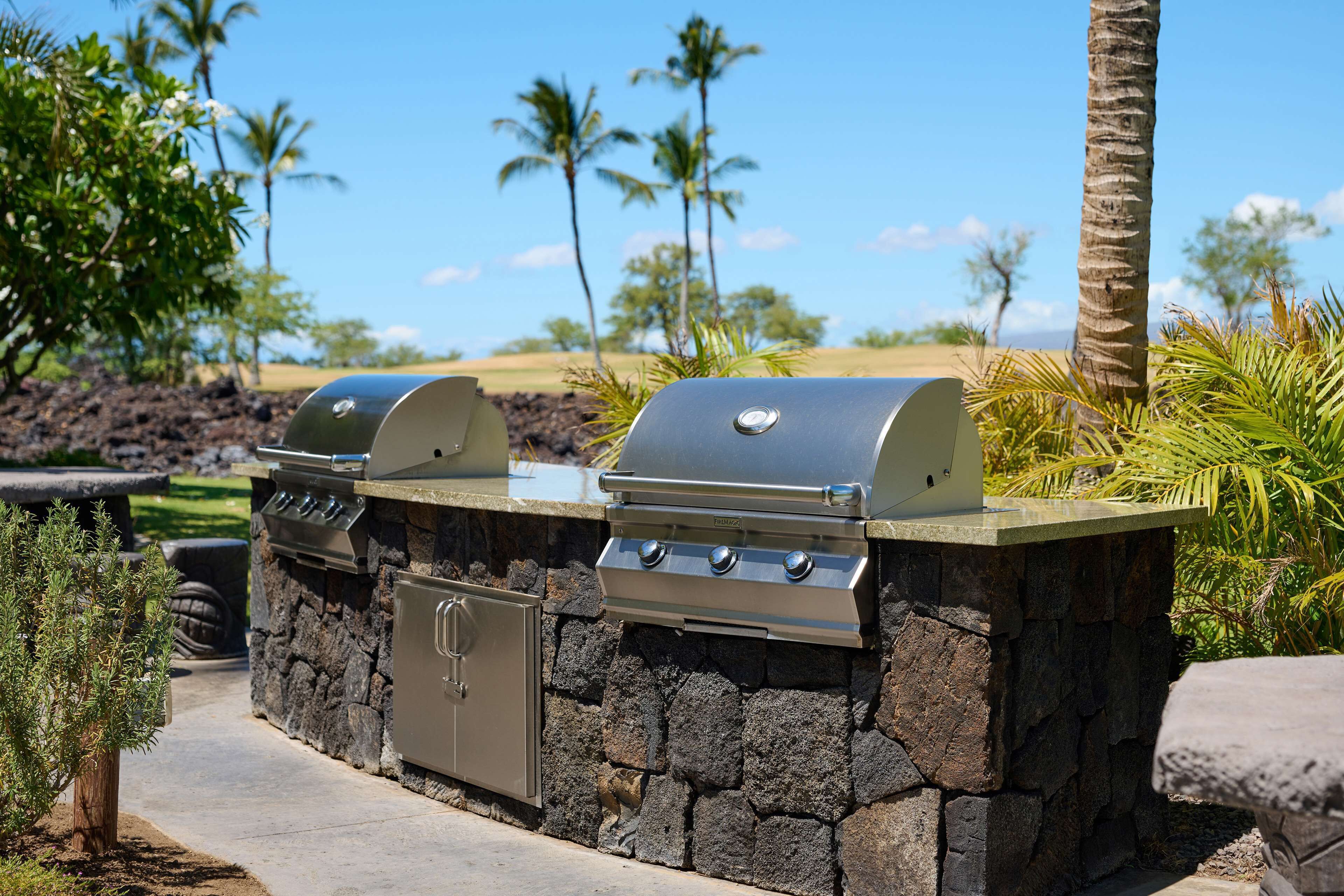 A stone barbecue grill with a built-in cooler and a side cabinet, set against a backdrop of palm trees and a clear sky.