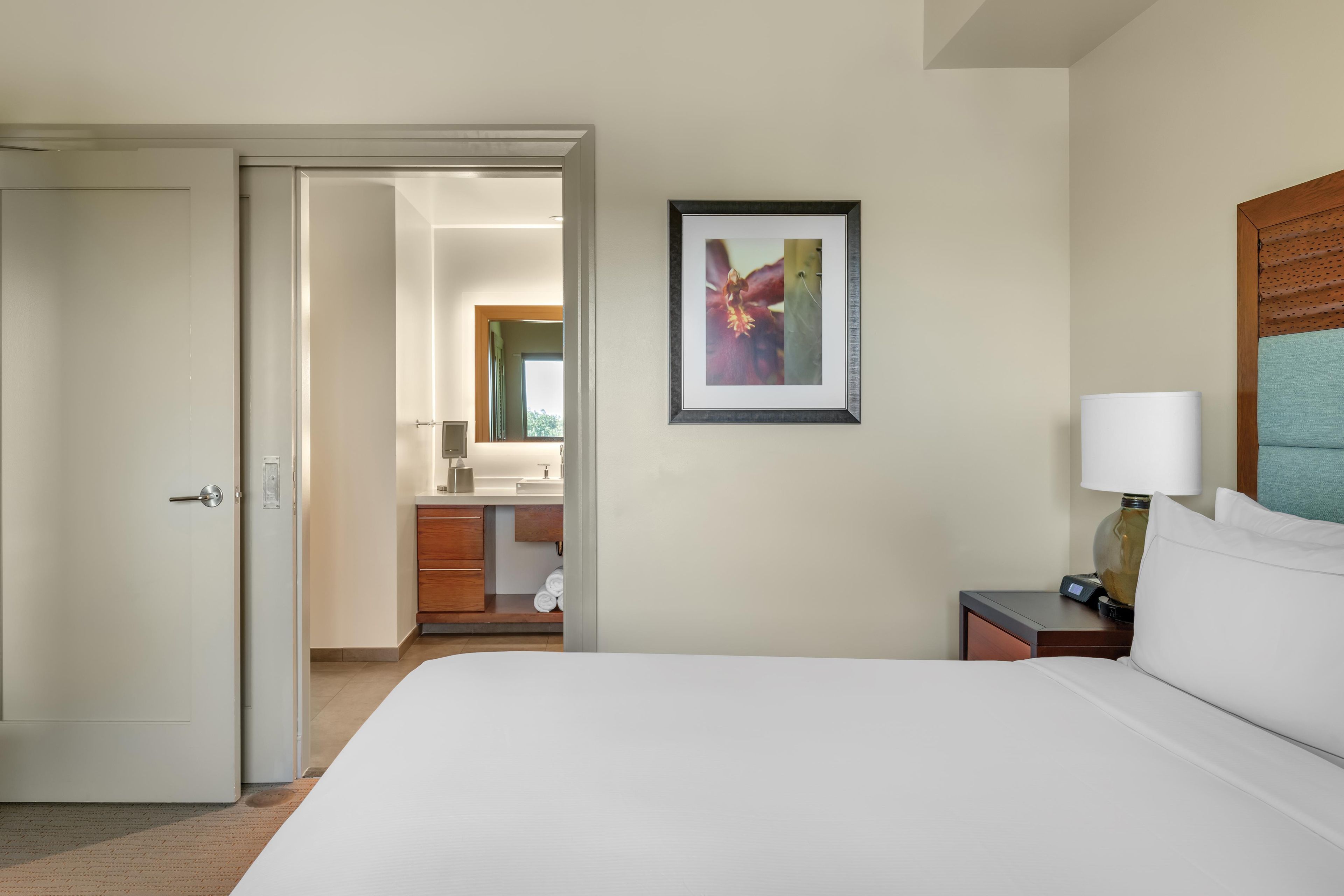 A hotel room with a bed, a nightstand with a lamp, a framed picture on the wall, and a bathroom visible through an open door.