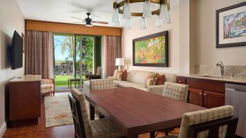 A spacious living area with a wooden dining table, comfortable seating, a ceiling fan, and a view of a lush garden through large glass doors.
