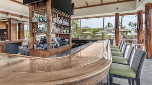 Un espace bar doté d’un comptoir incurvé, d’étagères en bois où sont exposées des bouteilles, et d’un coin salon extérieur avec des coussins verts.