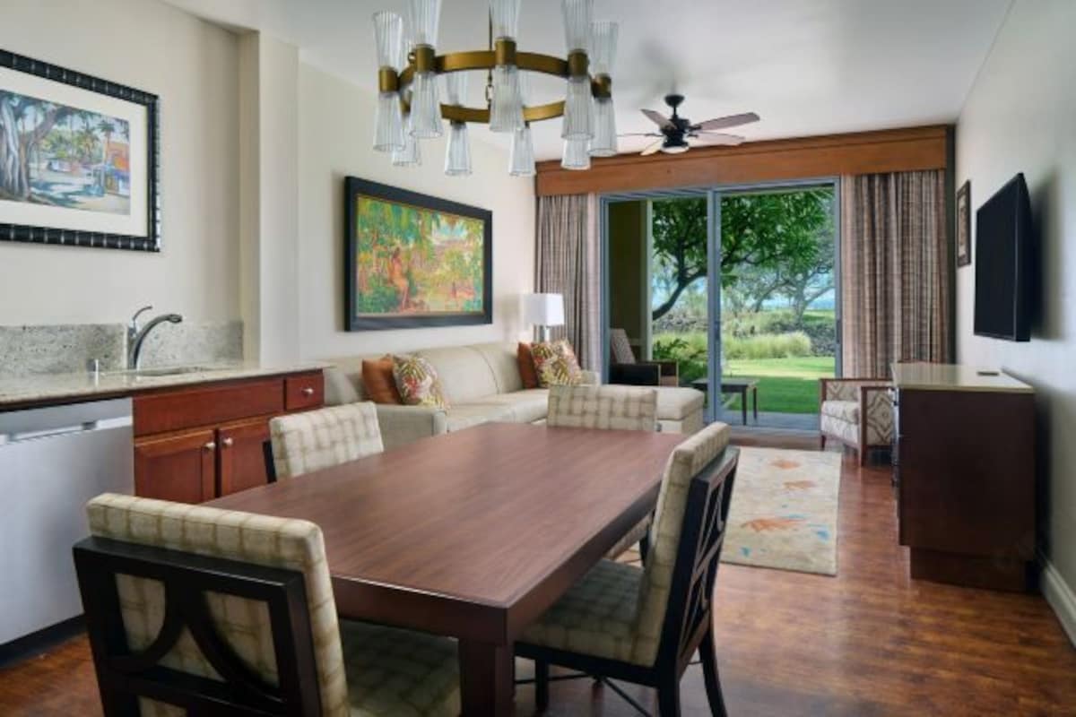 A spacious living area with a dining table, sofa, and chairs. There is a kitchen area with a sink and cabinets. A large window provides a view of the outdoors.