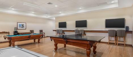 A room with pool tables, a ping pong table, and two flat-screen TVs mounted on the wall.