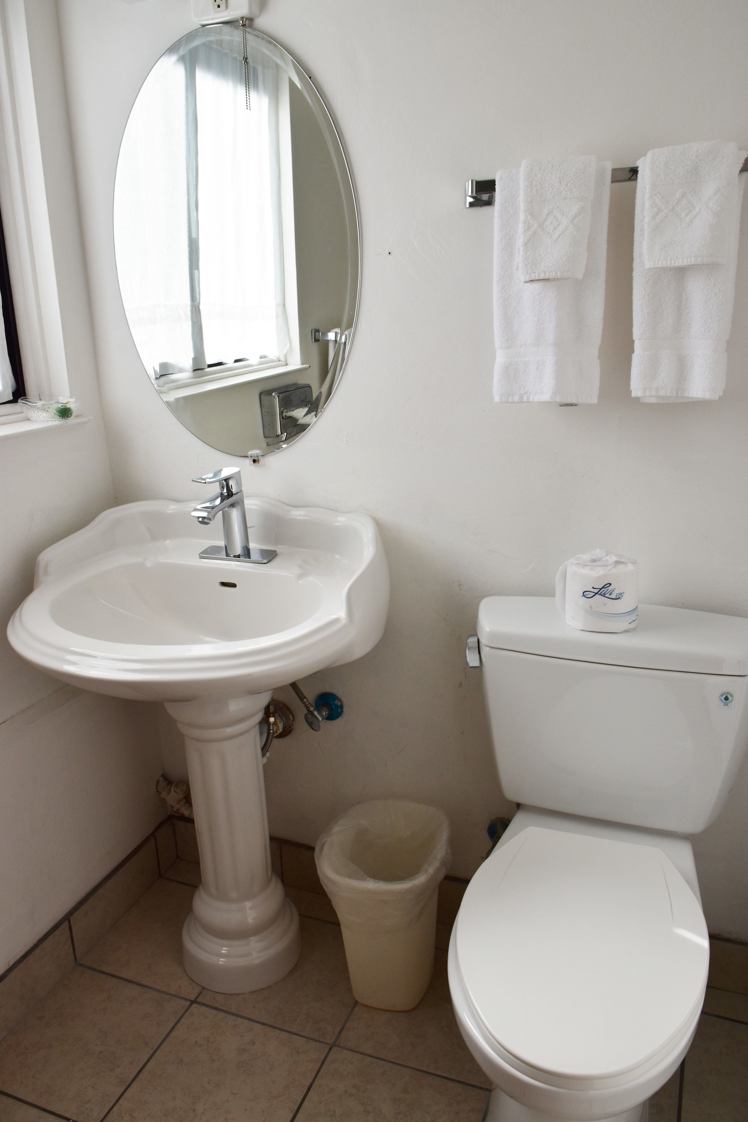 A bathroom with a pedestal sink, a toilet, a mirror, and a trash bin.