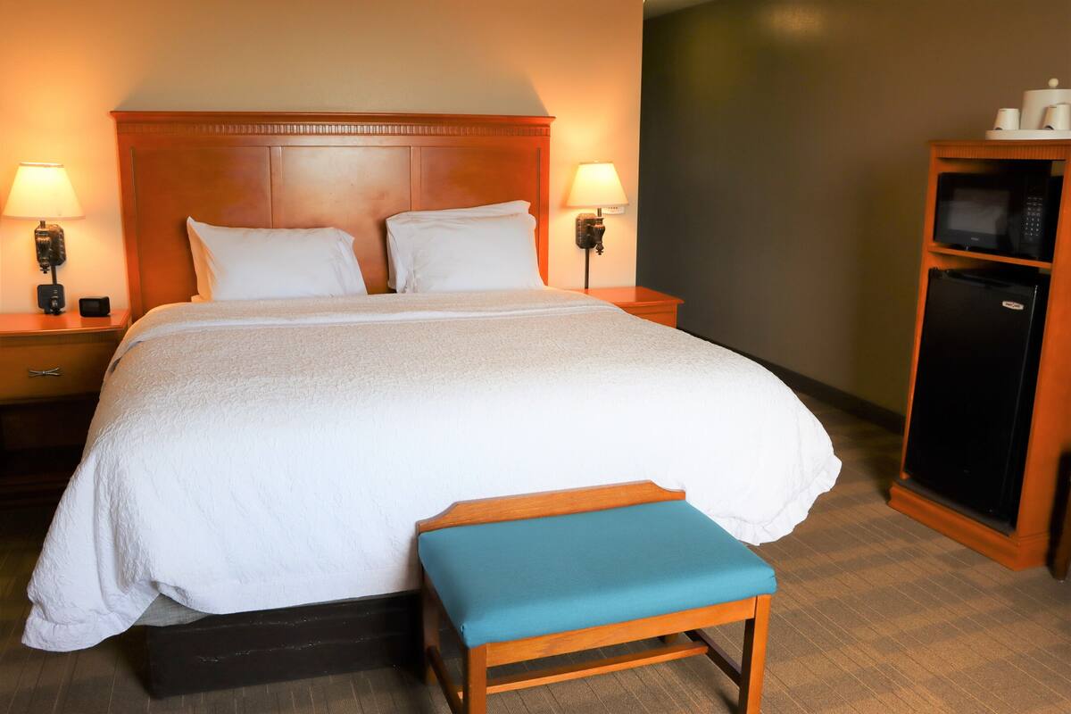 A neatly made bed with white linens, a wooden headboard, two bedside lamps, a small wooden bench, and a black refrigerator.