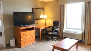 A hotel room with a flat-screen TV, a desk with a lamp, a chair, a wooden table, and a window with curtains.