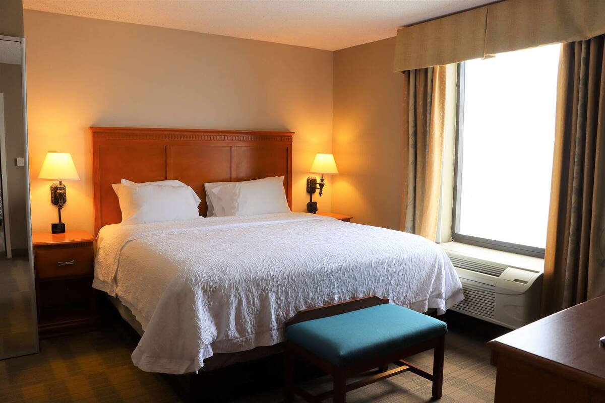 A hotel room with a large bed, two bedside lamps, a window with curtains, a desk, and a bench.