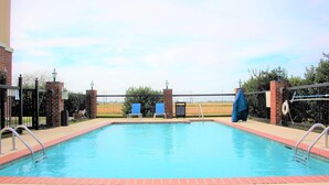 A rectangular swimming pool with a brick border, surrounded by a metal fence with a brick pillar and a lamp post. There are two blue chairs and a lifebuoy on the fence.