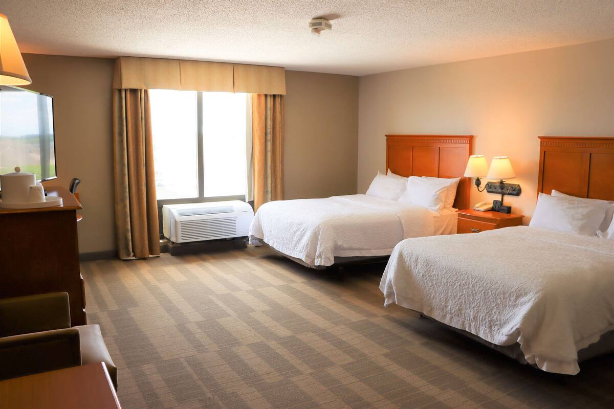 A hotel room with two beds, a desk, a chair, a lamp, and a window with curtains.