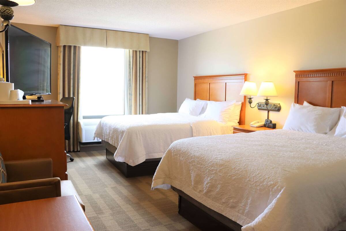 A hotel room with two beds, a desk, a chair, a TV, and a window with curtains.