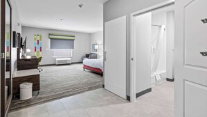 A modern hotel room with a bed, a desk, a chair, a trash bin, and a shower area.