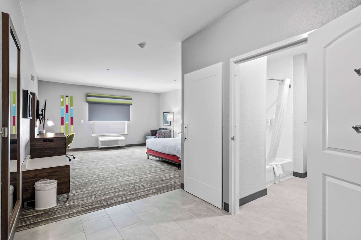 A modern hotel room with a bed, a desk, a chair, a trash bin, and a shower area.