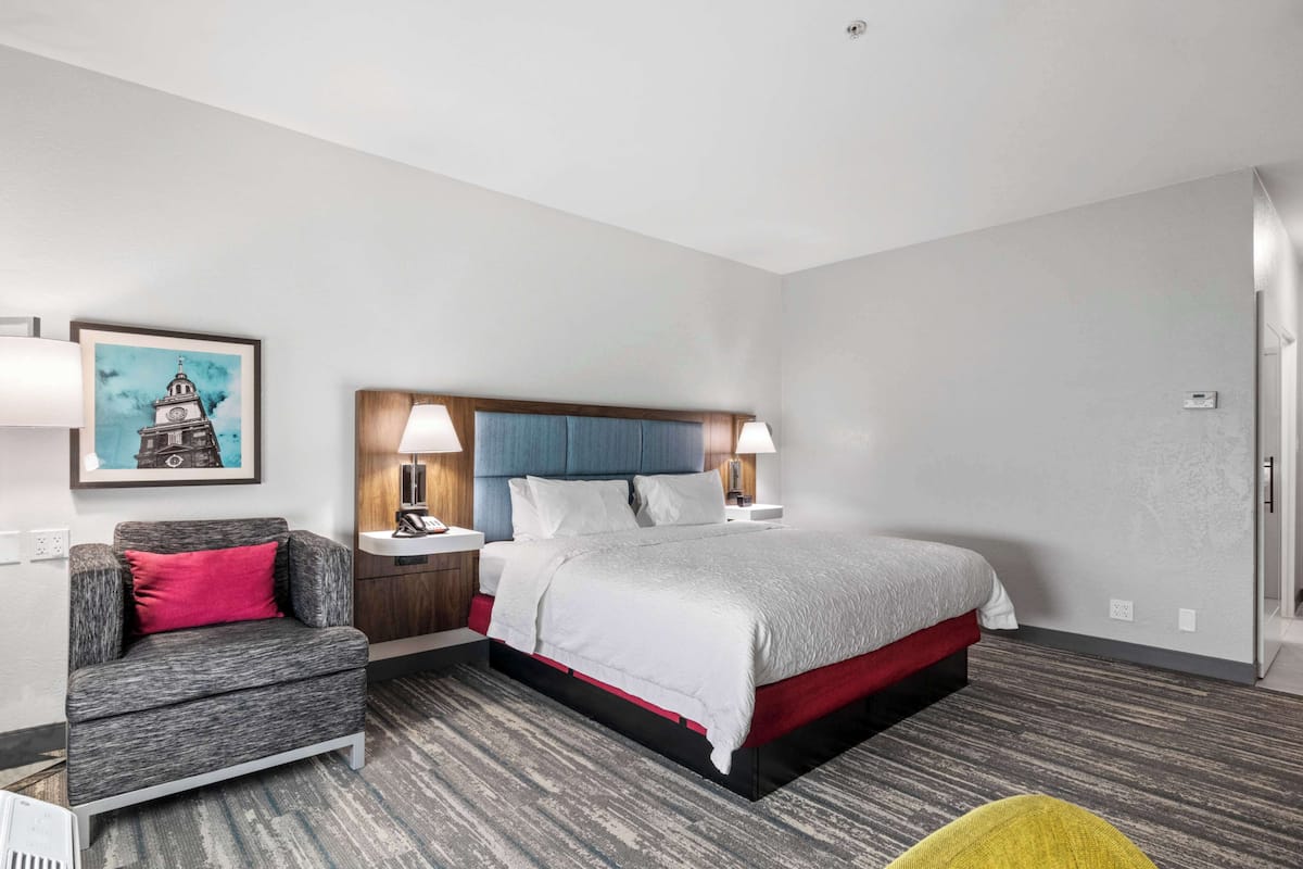 A hotel room with a bed, a chair, a nightstand, and a painting on the wall.