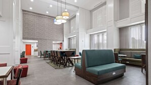 A spacious hotel lobby with a high ceiling, a dining area with tables and chairs, a seating area with a sofa, and a brick accent wall.