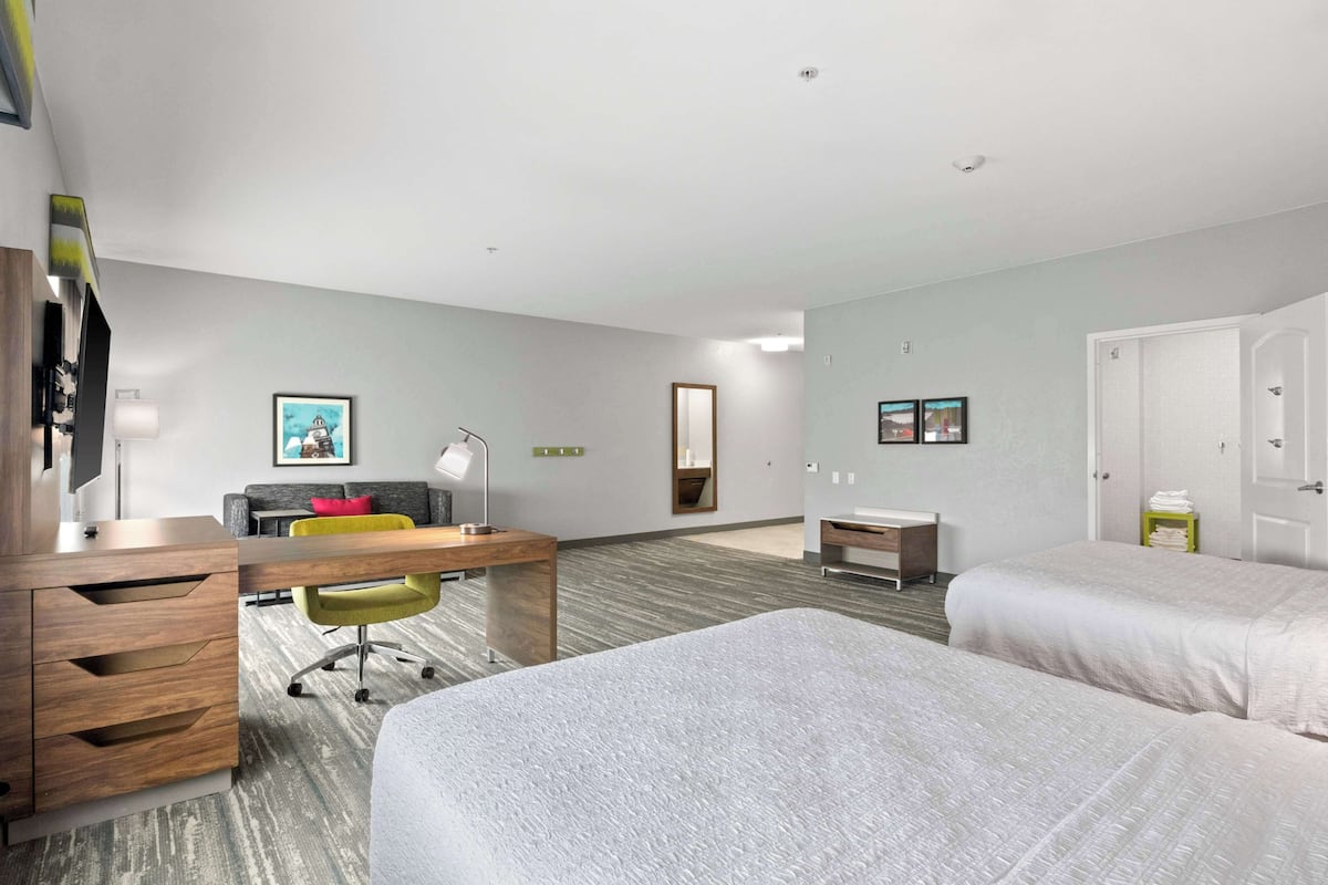 A hotel room with two beds, a desk, a chair, a small table, a mirror, and a TV.