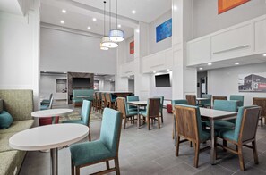 A modern dining area with round tables and teal chairs, a flat-screen TV mounted on the wall, and pendant lighting. - Hampton Inn & Suites Mount Pleasant (Mount Pleasant)
