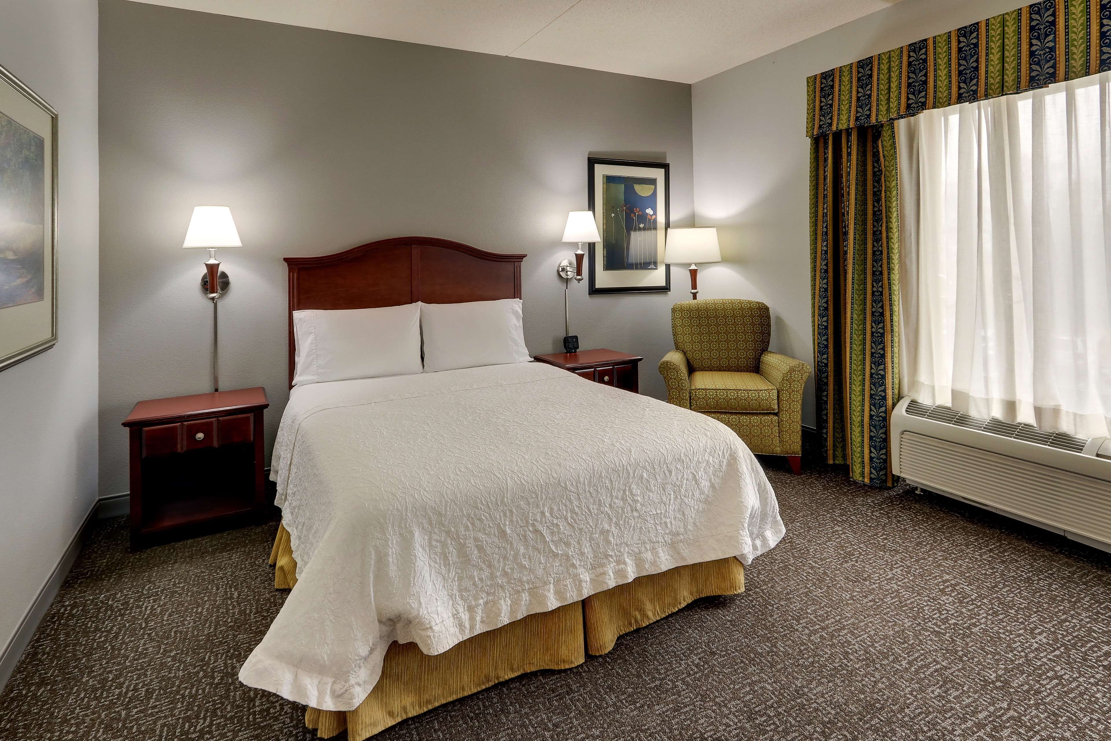 A hotel room with a large bed, two bedside tables, a chair, a framed picture on the wall, and a window with curtains.