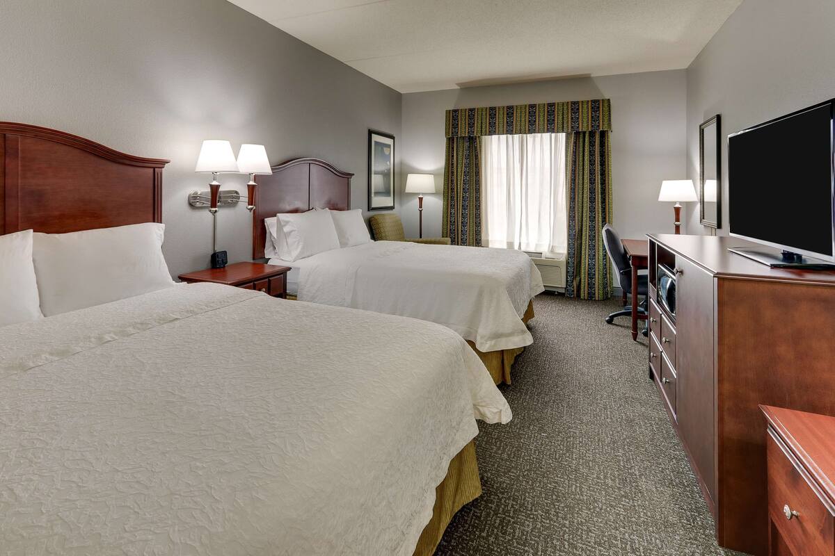 A hotel room with two beds, a desk, a television, and a window with curtains.