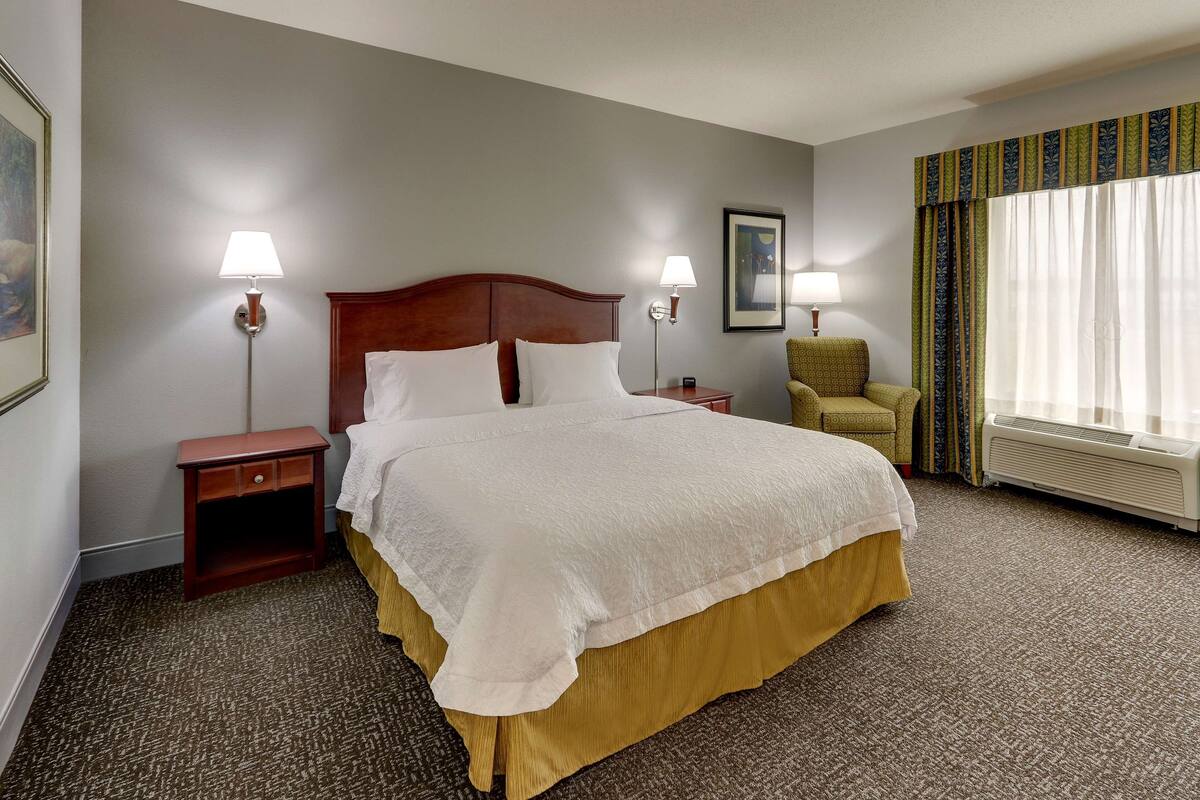 A hotel room with a large bed, two bedside tables with lamps, a chair, a window with curtains, and a framed picture on the wall.