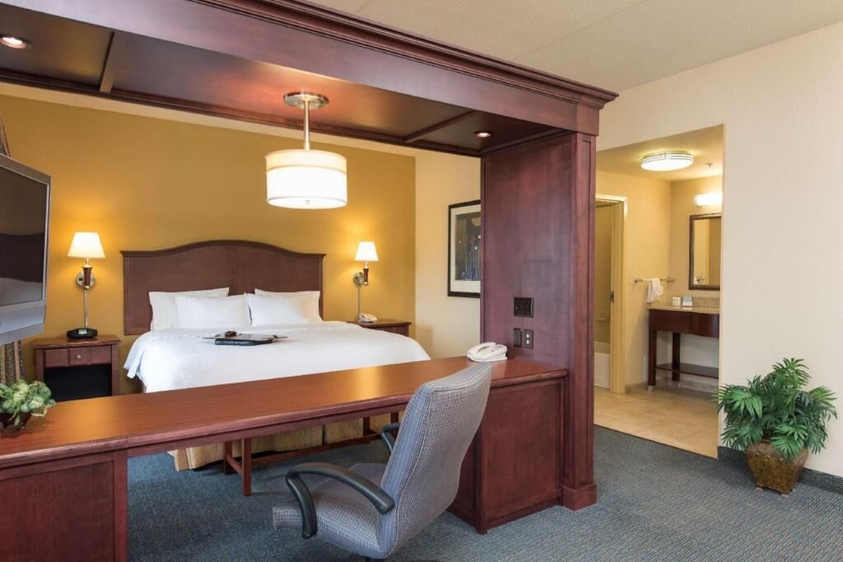 A hotel room with a bed, desk, chair, desk lamp, bedside table, and a potted plant.