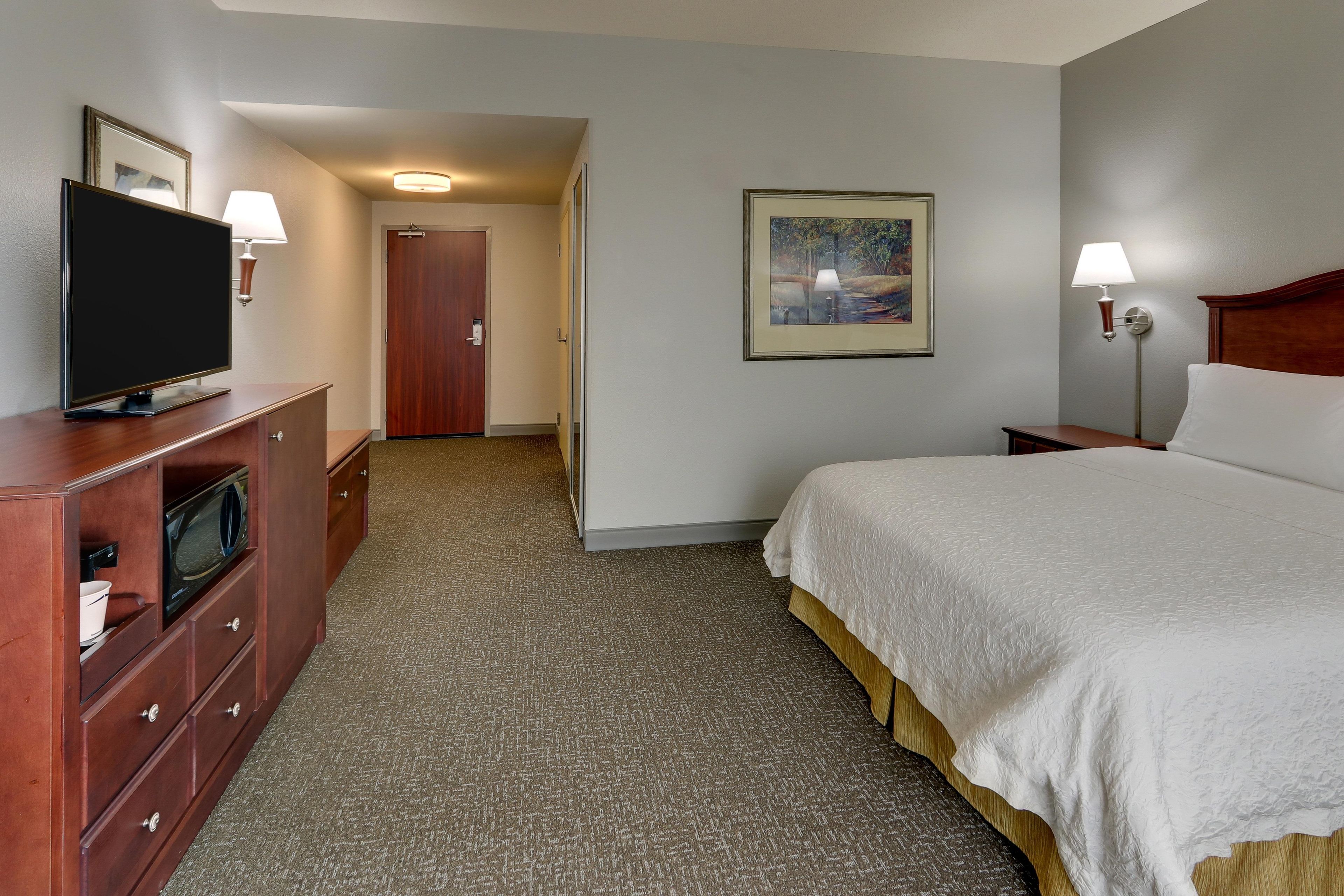 Habitación de hotel con una cama, un televisor en una cómoda, un cuadro en la pared y una puerta que da a otra habitación.