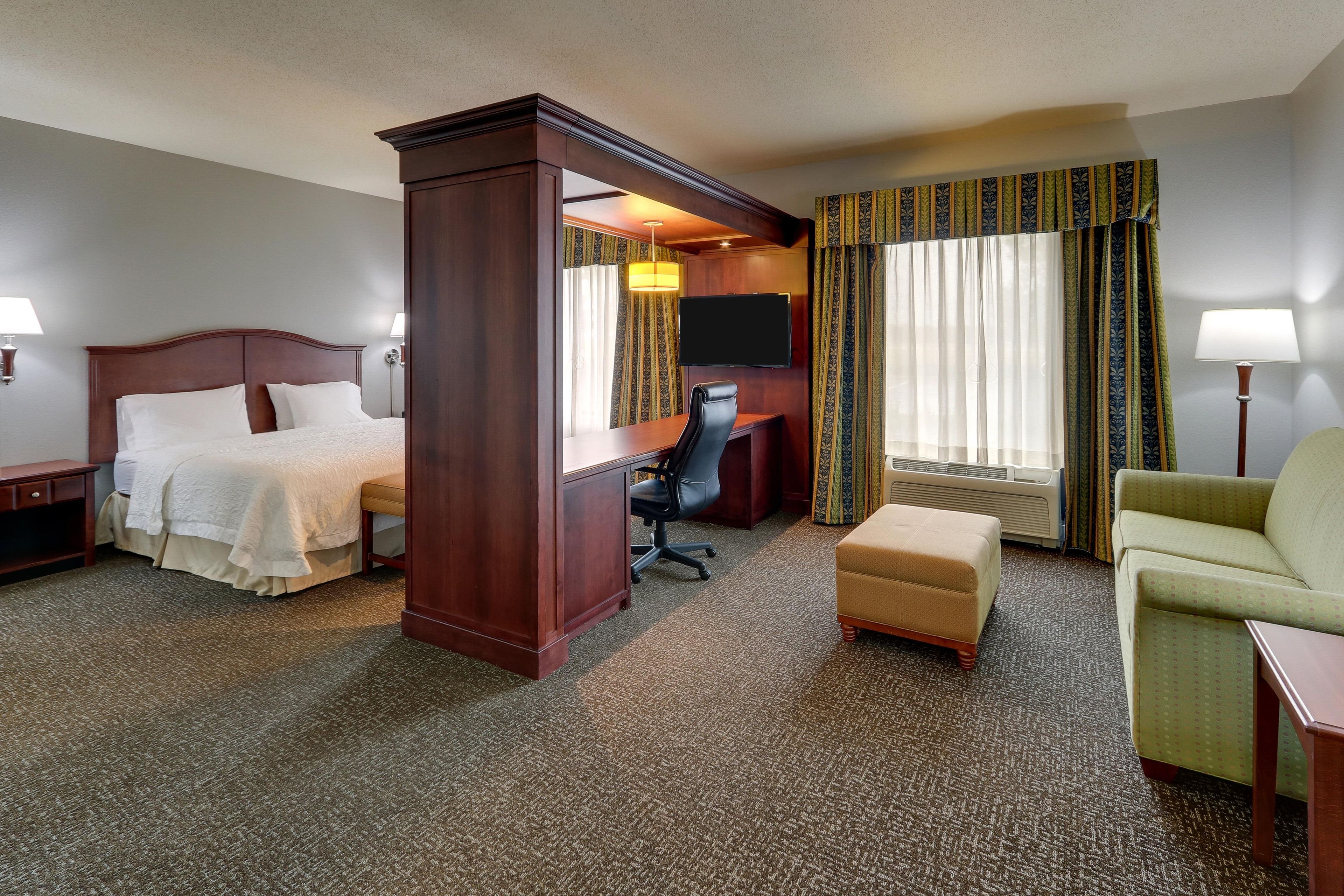 A hotel room with a king-sized bed, a desk, a chair, an ottoman, and a television.