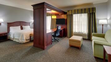 A hotel room with a king-sized bed, a desk, a chair, an ottoman, and a television.