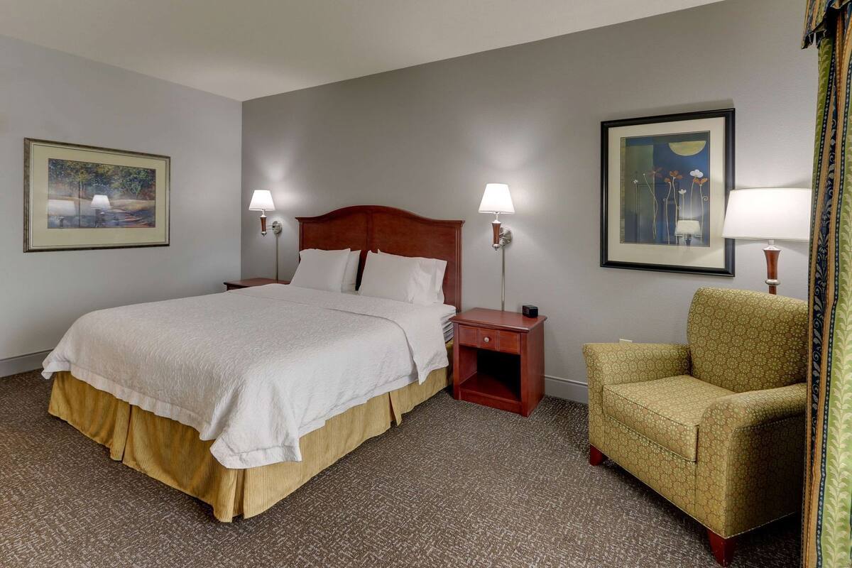 A hotel room with a large bed, two bedside tables with lamps, a chair, and two framed pictures on the walls.