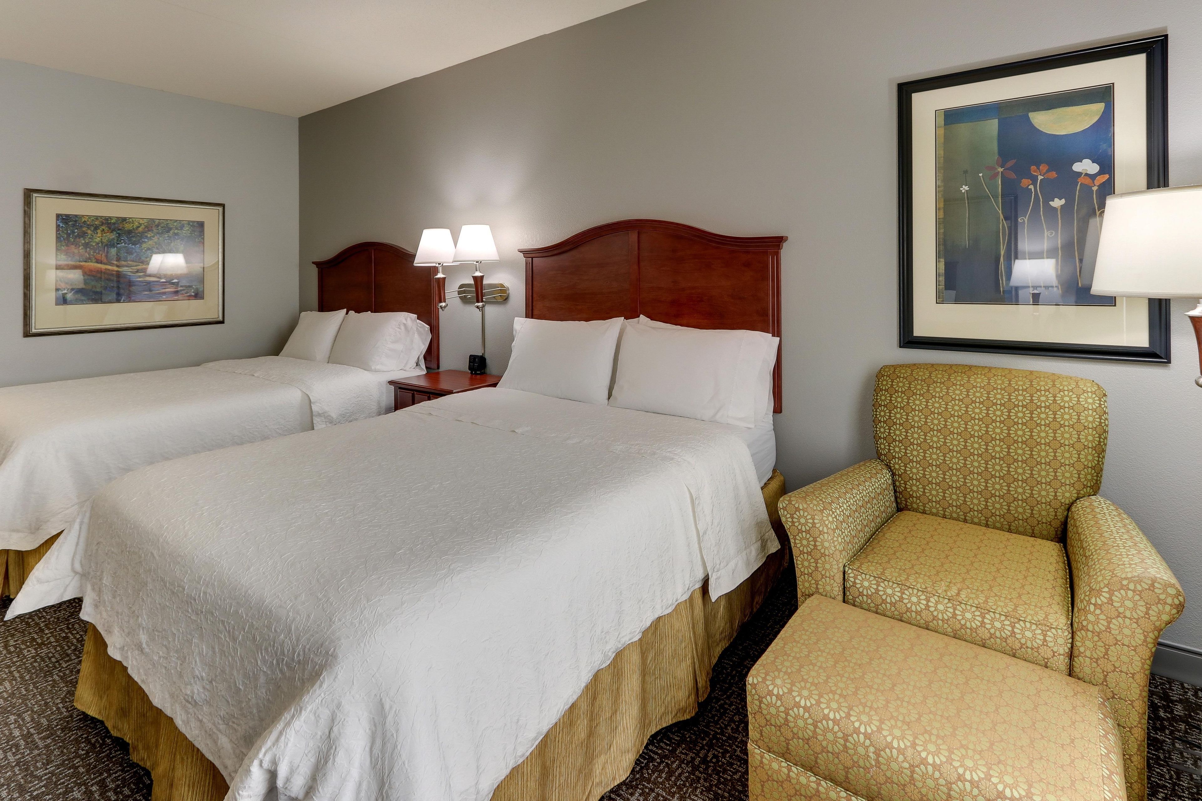 A hotel room with two beds, a nightstand, a lamp, and a chair.