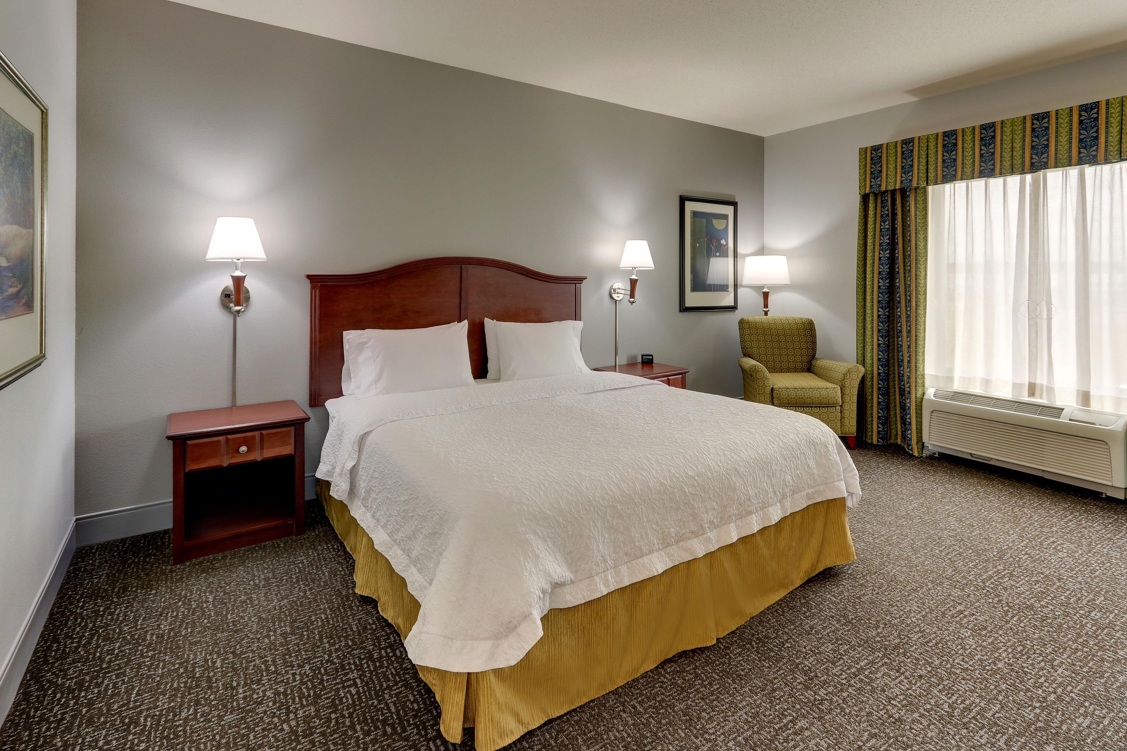 A hotel room with a large bed, two bedside tables, a chair, a painting, and a window with curtains.