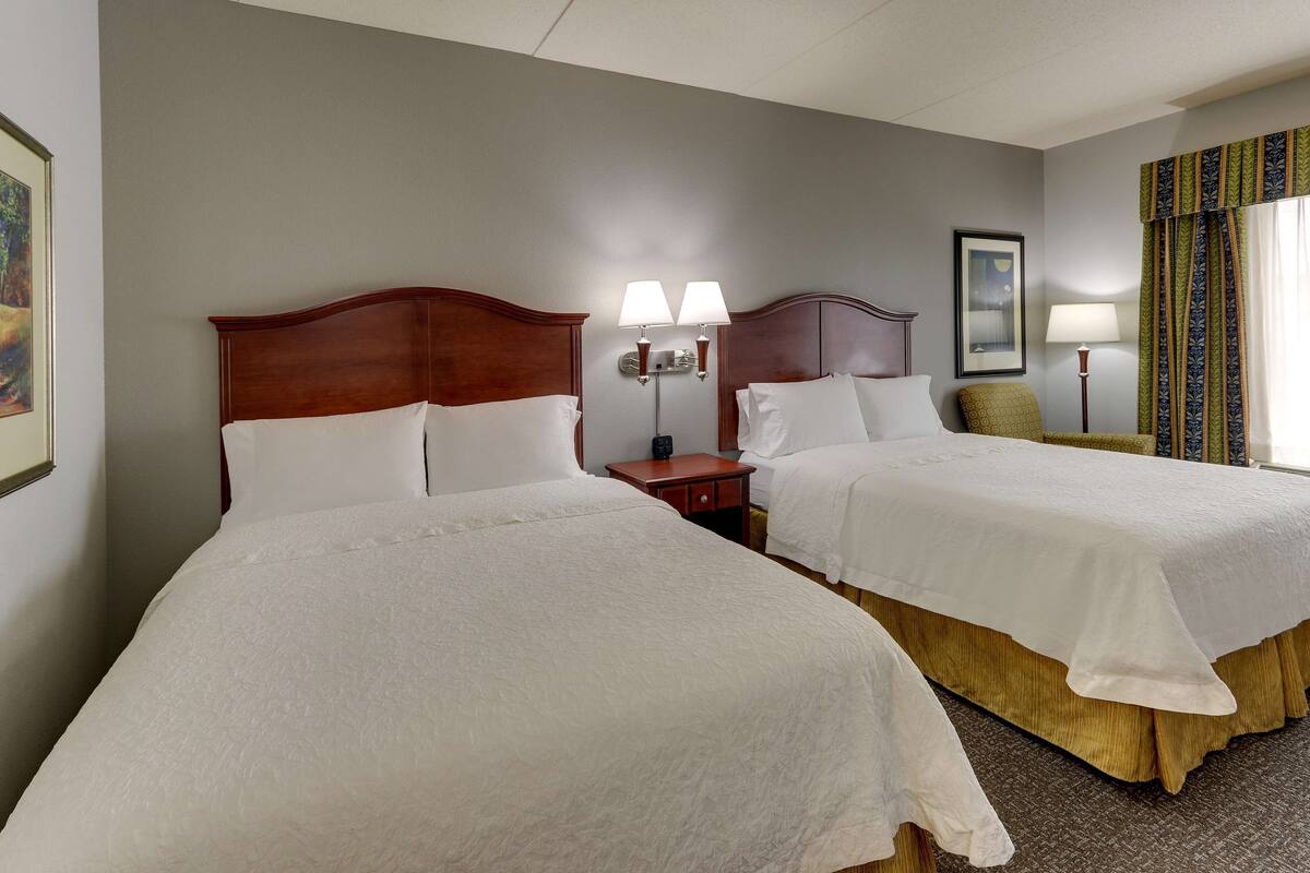 A hotel room with two beds, a nightstand, a lamp, and a framed picture on the wall.