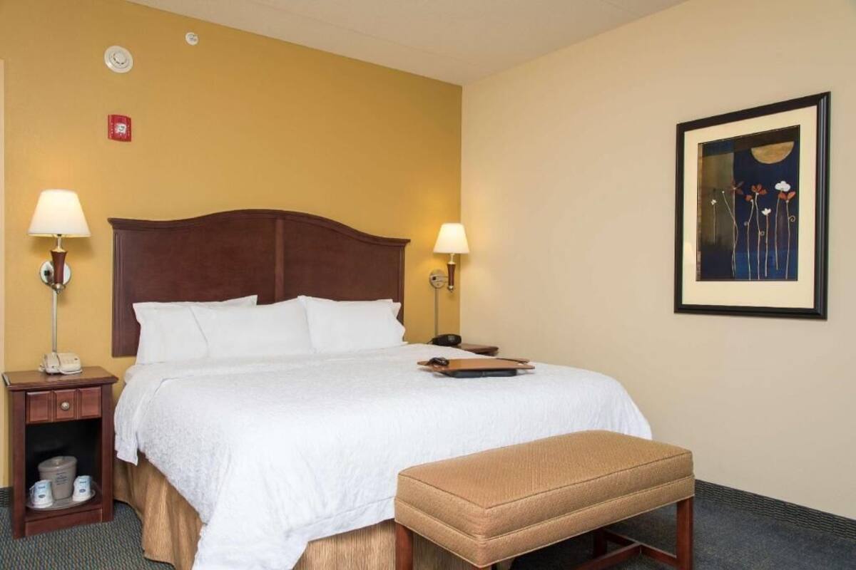A hotel room with a bed, bedside lamps, a framed artwork, and a small table with a phone and a bucket.